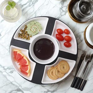 Puede incluir: Una fuente para servir dividida en blanco y negro con un cuenco central para mojar. La fuente contiene varios aperitivos, como tomates, galletas y un pastel verde. La fuente está colocada sobre una superficie de mármol, con tenedores y un vaso de agua visibles.