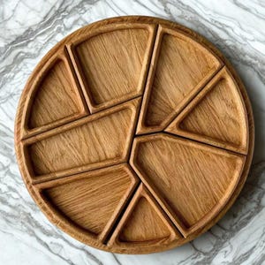 Op de afbeelding: Een ronde houten serveerschaal met meerdere compartimenten. De lichtbruine houten schaal is verdeeld in segmenten, ideaal voor het presenteren van hapjes of voorgerechten. De compartimenten hebben verschillende geometrische vormen.