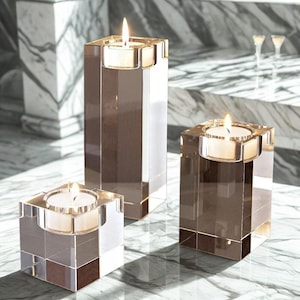 Puede incluir: Tres candelabros de vidrio transparente de diferentes alturas, cada uno con una vela blanca encendida. Los candelabros tienen un diseño cuadrado y rectangular, con un patrón geométrico reflectante. El fondo es una superficie de mármol.