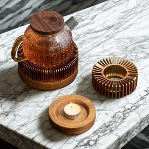 May include: A glass teapot with a wooden lid sits on a copper-colored warmer. A matching candle holder and a wooden coaster with a lit tea light are also present. The items are arranged on a marble surface.