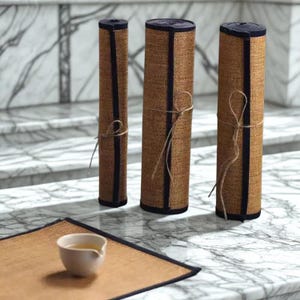 Puede incluir: Tres manteles individuales enrollados de tejido marrón con ribete azul marino, atados con hilo. Un cuarto mantel individual está plano con una pequeña taza de té blanca encima. Los manteles individuales están sobre una superficie de mármol.