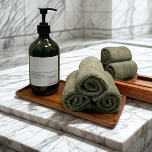 以下が含まれることがあります： 黒いポンプが付いた濃い緑色の石鹸ディスペンサーが、丸められた緑色のタオルと一緒に木製のトレイの上に置かれています。タオルは木製のトレイに配置されています。背景には大理石の表面があります。