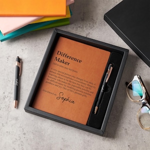May include: A black gift set featuring a tan notebook and a pen. The notebook has the words "Difference Maker" and a definition. The black pen has gold detailing and the name "Sophia". The set is presented in a black box.