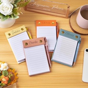 Puede incluir: Cuatro blocs de notas rectangulares de imitación cuero en amarillo, naranja, rosa y azul, cada uno con un nombre personalizado, remaches metálicos dorados y un bolígrafo a juego. Los blocs tienen papel blanco rayado sobre madera clara.