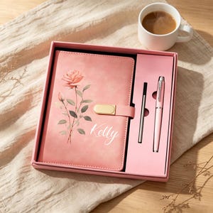 May include: A pink gift set containing a rectangular notebook with a rose illustration and the name Kelly printed in white, a matching pink pen, and a black ink refill, all housed in a pink box with a gold-toned metal magnetic closure.
