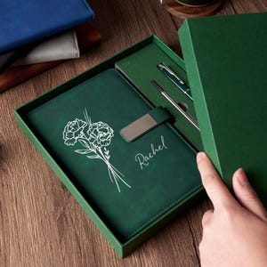 Peut inclure: Un ensemble de journal et de stylo en cuir vert dans une boîte cadeau. Le journal présente un motif floral et le nom "Rachel" en écriture blanche. Un stylo et un stylet argentés sont inclus.