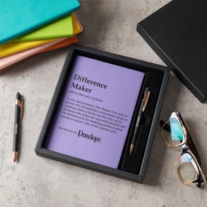 May include: A black gift set featuring a pen and a notepad with the definition of "Difference Maker". The pen has rose gold accents and the notepad is lavender. A pair of glasses and other stationery are also visible.