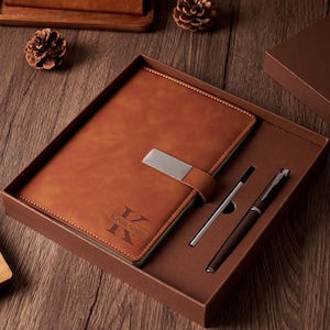 May include: A brown leather journal and pen set in a gift box. The journal has a silver clasp and the name "Kevin Smith" engraved on the cover. Two pens, one silver and one brown, are included.