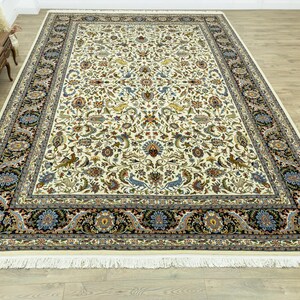 May include: A large, rectangular ivory-colored rug with an intricate floral pattern in shades of blue, brown, and green. The rug has a black border with floral and paisley designs, and a fringed edge. The rug is placed on a hardwood floor.