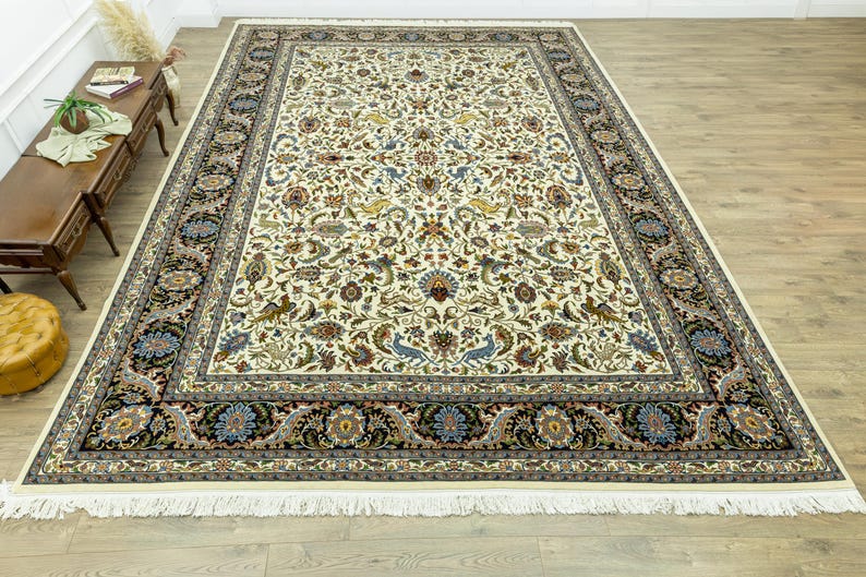 May include: A large, rectangular ivory-colored rug with an intricate floral pattern in shades of blue, brown, and green. The rug has a black border with floral and paisley designs, and a fringed edge. The rug is placed on a hardwood floor.