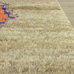 Peut inclure: Gros plan sur un tapis beige et touffu avec un motif circulaire orange et bleu dans le coin sup&eacute;rieur gauche. Le tapis a un poil &eacute;pais et textur&eacute;, et le fond est un plancher en bois clair.