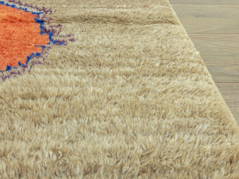 Peut inclure: Gros plan sur un tapis beige et touffu avec un motif circulaire orange et bleu dans le coin sup&eacute;rieur gauche. Le tapis a un poil &eacute;pais et textur&eacute;, et le fond est un plancher en bois clair.