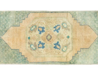 May include: Rectangular rug with a central geometric design in beige, blue, and green. The rug has a light beige center with blue floral motifs and a green border. The rug is likely made of wool or a similar material.