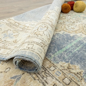 May include: Partially rolled vintage-style rug with a distressed, faded appearance. The rug features a mix of beige, blue, and brown tones with intricate patterns. Three balls of yarn in orange and yellow sit on the rug.