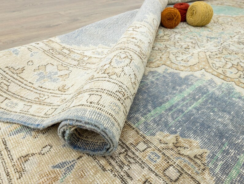 May include: Partially rolled vintage-style rug with a distressed, faded appearance. The rug features a mix of beige, blue, and brown tones with intricate patterns. Three balls of yarn in orange and yellow sit on the rug.