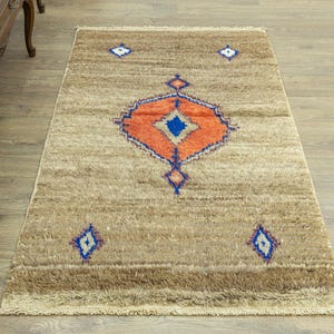 Peut inclure: Tapis rectangulaire beige avec un motif g&eacute;om&eacute;trique central orange, bleu et blanc. Le tapis pr&eacute;sente un motif de losanges bleu et blanc r&eacute;p&eacute;t&eacute;. Le tapis a une bordure frang&eacute;e et convient &agrave; la d&eacute;coration int&eacute;rieure.