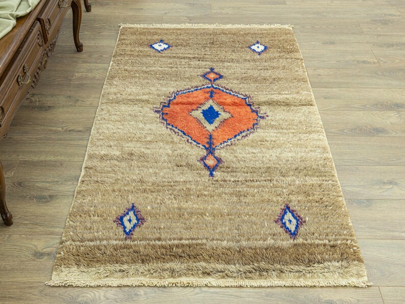 Peut inclure: Tapis rectangulaire beige avec un motif g&eacute;om&eacute;trique central orange, bleu et blanc. Le tapis pr&eacute;sente un motif de losanges bleu et blanc r&eacute;p&eacute;t&eacute;. Le tapis a une bordure frang&eacute;e et convient &agrave; la d&eacute;coration int&eacute;rieure.