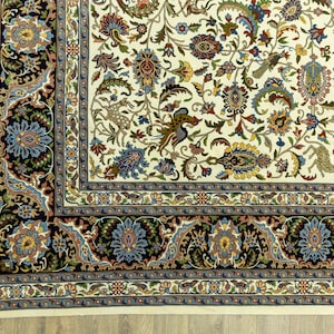 May include: Close-up of an ornate, cream-colored rug with a detailed floral pattern. The rug features a black border with blue and gold floral designs, and a cream fringe along one edge. The central design includes various flowers and animal figures.