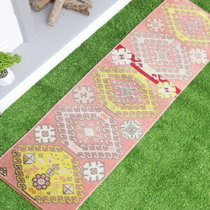 May include: A pink and yellow patterned runner rug with a geometric design. The rug is on a green artificial grass surface.