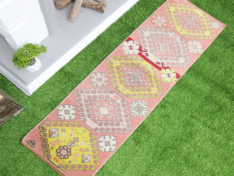 May include: A pink and yellow patterned runner rug with a geometric design. The rug is on a green artificial grass surface.