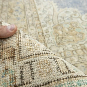 May include: Close-up of a rolled-up, vintage-style rug. The rug features a beige and light blue color palette with intricate patterns and a distressed look. The edges show a detailed border with geometric designs.
