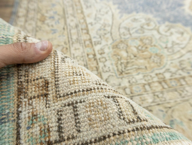 May include: Close-up of a rolled-up, vintage-style rug. The rug features a beige and light blue color palette with intricate patterns and a distressed look. The edges show a detailed border with geometric designs.