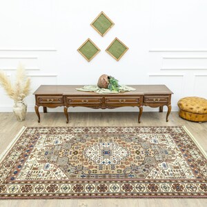 May include: A beige rug with a brown, cream, and blue patterned design. The rug is laid out on a wooden floor in front of a wooden console table with drawers. A brown leather ottoman is on the right side of the image.