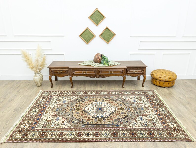 May include: A beige rug with a brown, cream, and blue patterned design. The rug is laid out on a wooden floor in front of a wooden console table with drawers. A brown leather ottoman is on the right side of the image.