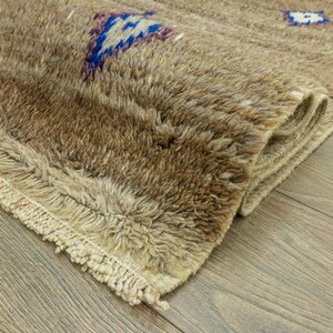 Peut inclure: Gros plan sur un tapis beige textur&eacute; avec un motif g&eacute;om&eacute;trique bleu et violet. Le tapis a un poil touffu et un bord &agrave; franges. Le tapis est partiellement enroul&eacute;, r&eacute;v&eacute;lant son envers.
