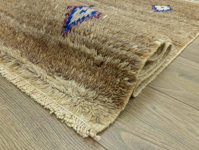 Peut inclure: Gros plan sur un tapis beige textur&eacute; avec un motif g&eacute;om&eacute;trique bleu et violet. Le tapis a un poil touffu et un bord &agrave; franges. Le tapis est partiellement enroul&eacute;, r&eacute;v&eacute;lant son envers.