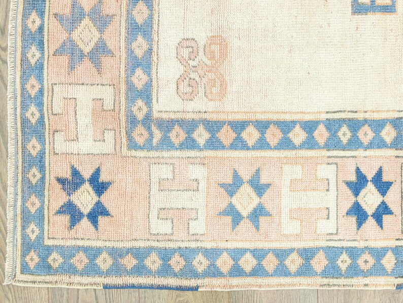 May include: Close-up of a beige rug with a blue and white geometric pattern. The rug has a fringed border and features a repeating star design.