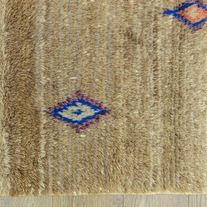 Peut inclure: Gros plan sur un tapis beige textur&eacute; avec un motif g&eacute;om&eacute;trique. Le tapis pr&eacute;sente des formes de losanges bleus, violets et oranges. Le tapis a une frange sur un c&ocirc;t&eacute; et est pos&eacute; sur un sol en bois.