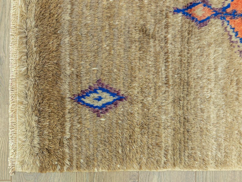Peut inclure: Gros plan sur un tapis beige textur&eacute; avec un motif g&eacute;om&eacute;trique. Le tapis pr&eacute;sente des formes de losanges bleus, violets et oranges. Le tapis a une frange sur un c&ocirc;t&eacute; et est pos&eacute; sur un sol en bois.