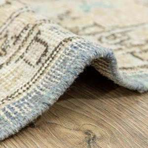 May include: Close-up of a rolled-up, vintage-style rug with a distressed pattern. The rug features a beige and light blue color palette with intricate geometric designs. The edges are finished with a light blue border, and the rug is placed on a wooden surface.