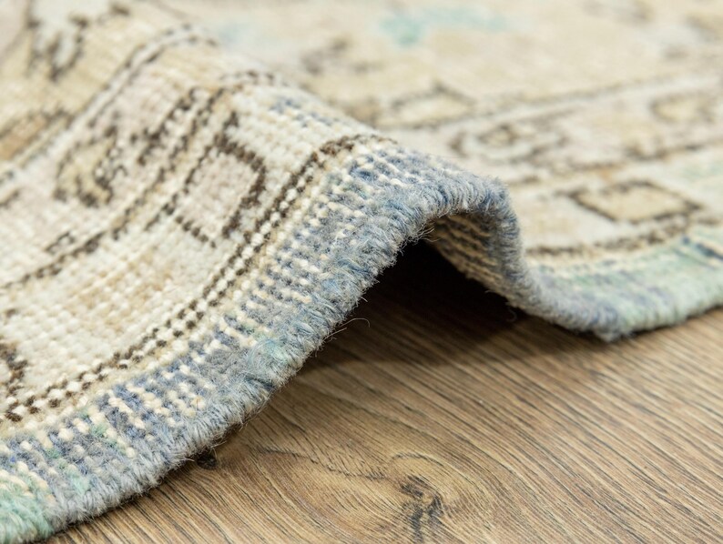 May include: Close-up of a rolled-up, vintage-style rug with a distressed pattern. The rug features a beige and light blue color palette with intricate geometric designs. The edges are finished with a light blue border, and the rug is placed on a wooden surface.