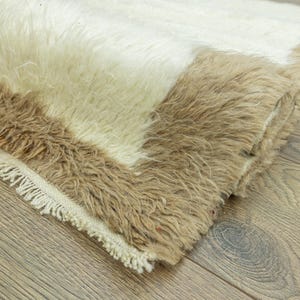 May include: A rectangular rug with a shaggy texture. The rug features a color block design with sections of white, beige, and brown. The edges are finished with a fringe.