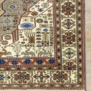 May include: Close-up of a cream-colored rug with a detailed geometric pattern in shades of brown, red, blue, and green. The rug has a fringe border.