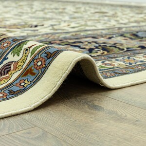 May include: Close-up of a rolled-up, cream-colored rug with an intricate floral and bird pattern. The border features a blue, green, and orange design. The rug is on a wooden floor.
