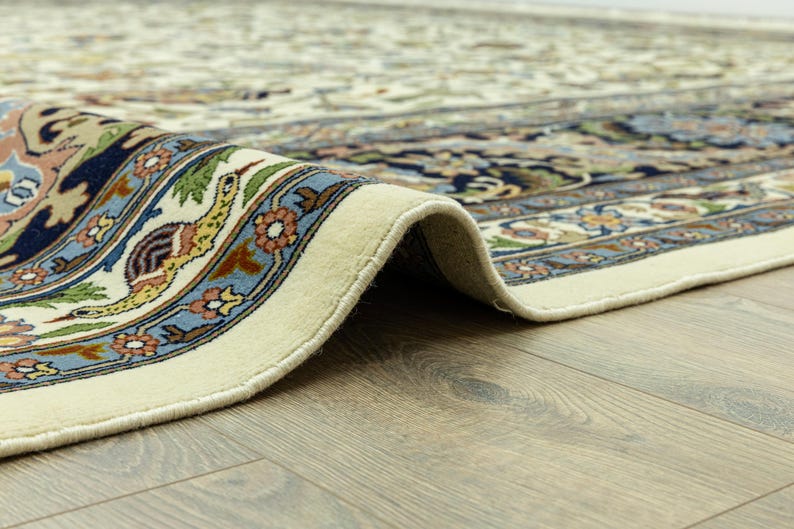 May include: Close-up of a rolled-up, cream-colored rug with an intricate floral and bird pattern. The border features a blue, green, and orange design. The rug is on a wooden floor.