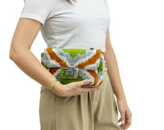 May include: A rectangular clutch with a colorful geometric pattern in orange, green, and gray. The bag has a flap closure with a silver-toned clasp. The clutch is held by a person wearing a white t-shirt and tan pants.