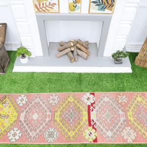 May include: A pink and yellow patterned runner rug with geometric designs. The rug is laid out on a green artificial grass surface in front of a fireplace.