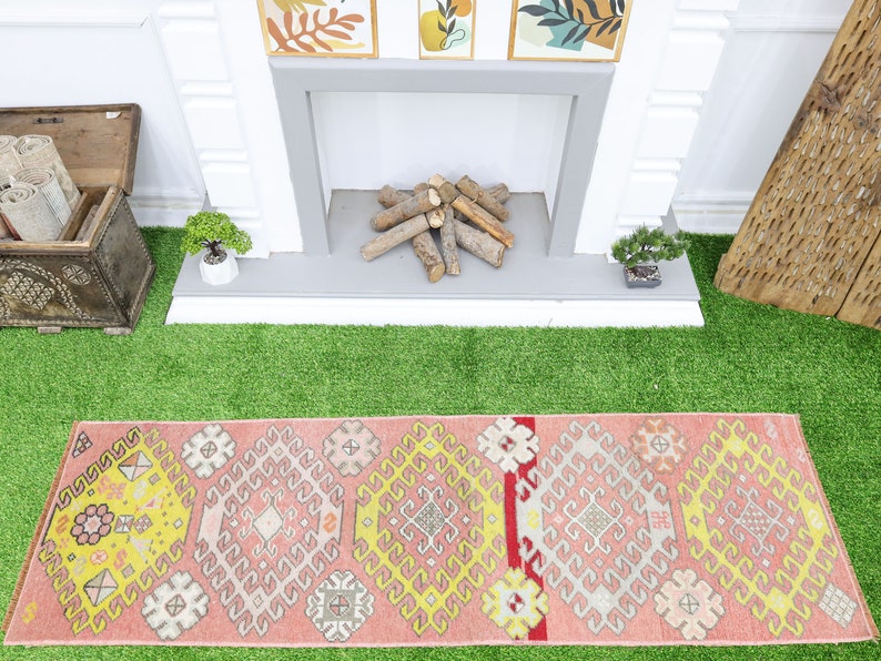 May include: A pink and yellow patterned runner rug with geometric designs. The rug is laid out on a green artificial grass surface in front of a fireplace.