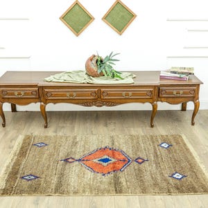 Peut inclure: Tapis rectangulaire marron avec un motif central en losange orange et bleu. Le tapis a une bordure effiloch&eacute;e et est pos&eacute; sur un sol en bois. Des objets d&eacute;coratifs sont sur une table en bois &agrave; l'arri&egrave;re-plan.
