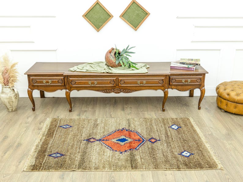 Peut inclure: Tapis rectangulaire marron avec un motif central en losange orange et bleu. Le tapis a une bordure effiloch&eacute;e et est pos&eacute; sur un sol en bois. Des objets d&eacute;coratifs sont sur une table en bois &agrave; l'arri&egrave;re-plan.