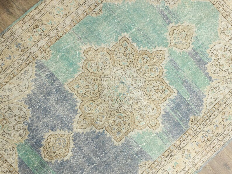 May include: A distressed, vintage-style rug with a central medallion design. The rug features a color palette of teal, beige, and muted blue. The border has an ornate pattern, and the rug has a faded, worn appearance.