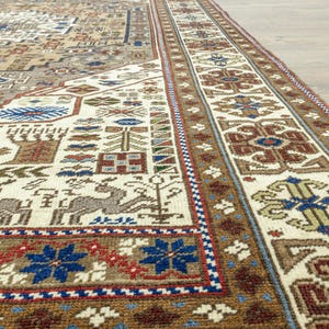 May include: Close-up of a beige and brown rug with intricate geometric patterns in shades of blue, red, and white. The rug has a fringe border.