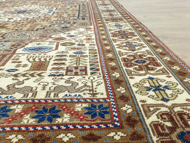May include: Close-up of a beige and brown rug with intricate geometric patterns in shades of blue, red, and white. The rug has a fringe border.