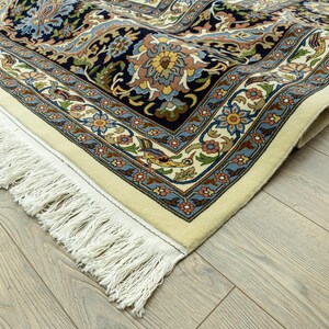 May include: Close-up of a Persian rug with a cream-colored border and intricate floral patterns in navy blue, beige, and light blue. The rug has a detailed design with a fringe edge.