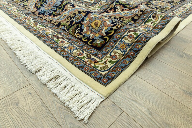 May include: Close-up of a Persian rug with a cream-colored border and intricate floral patterns in navy blue, beige, and light blue. The rug has a detailed design with a fringe edge.