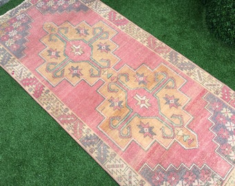 May include: A rectangular, vintage-style rug with a central design in shades of peach, tan, and teal. The rug features a geometric pattern with floral and star-like motifs, framed by a border of gray and beige. The rug is placed on a green surface.